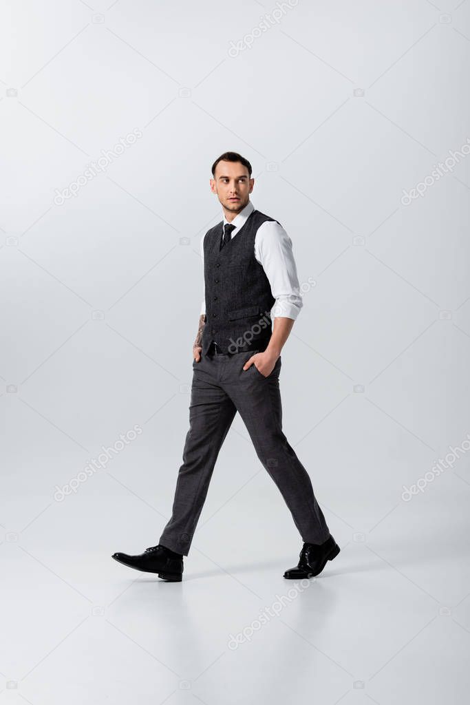 handsome tattooed elegant bridegroom walking with hands in pockets on grey background