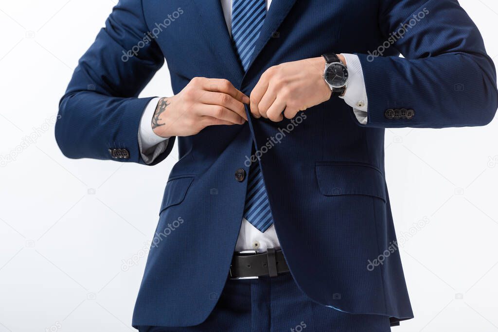 cropped view of successful tattooed young businessman in blue suit buttoning blazer isolated on white