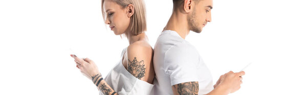 young tattooed couple using smartphones while standing back to back isolated on white