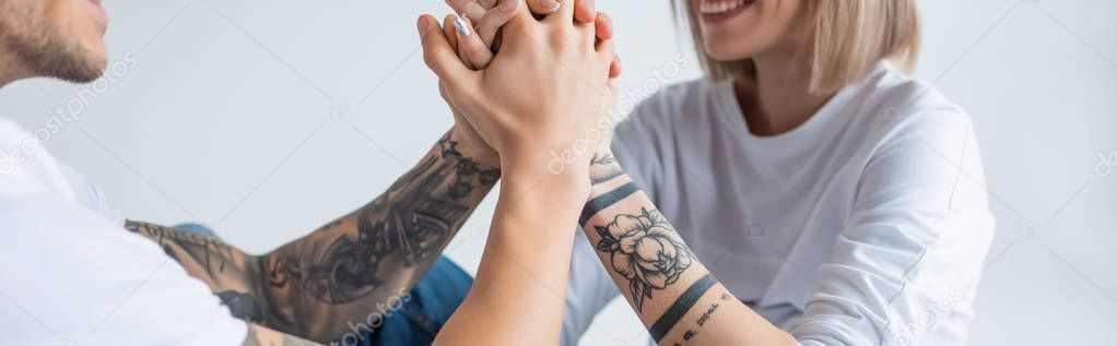 cropped view of smiling young tattooed woman sitting with husband on floor and holding hands isolated on grey, panoramic shot