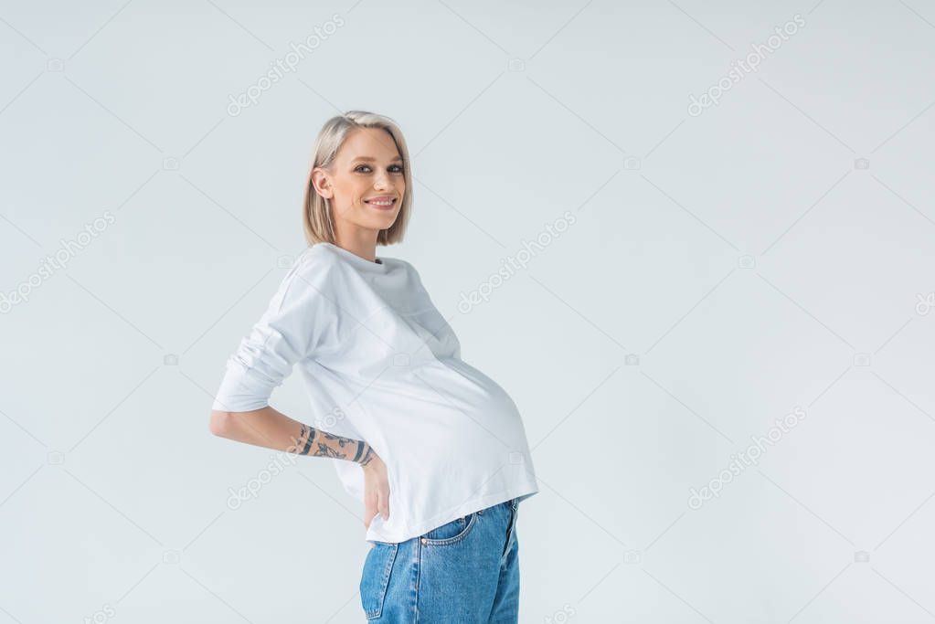 side view of happy young tattooed pregnant woman isolated on grey