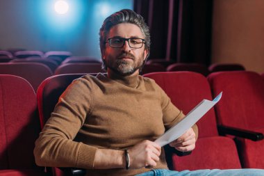 Tiyatrodaki koltuklarda senaryo yazan sakallı sahne yönetmeni. 