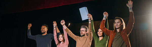 panoramic shot of happy actors and actresses with hands up on stage