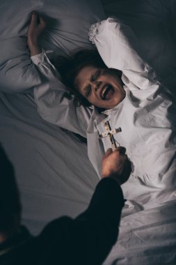 exorcist holding cross over creepy screaming demon in bed clipart