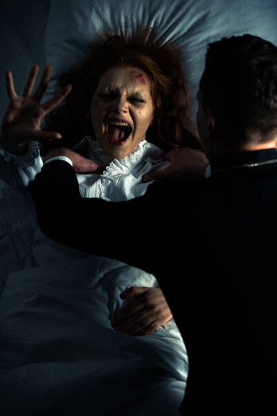 экзорцист держит кричащую одержимую девушку в постели
