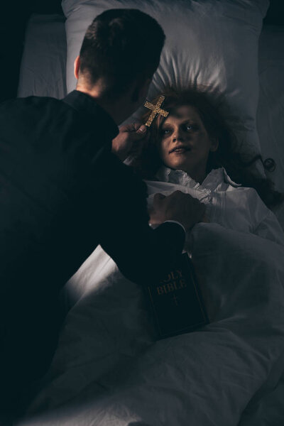 exorcist with bible and cross standing over obsessed girl in bed
