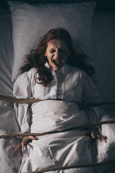 demonic obsessed yelling girl in nightgown bound with rope in bed