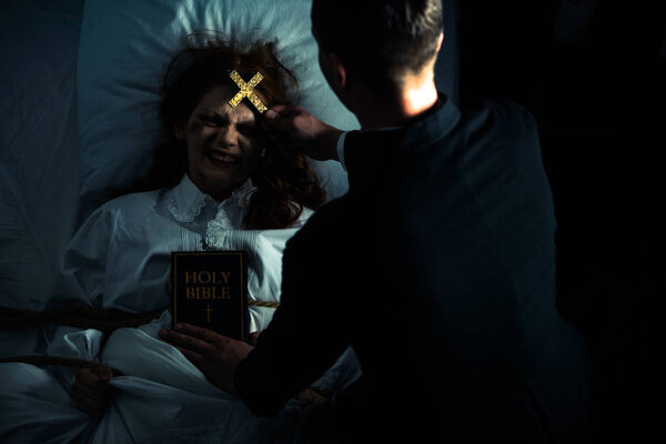 exorcist with bible and cross standing over demonic obsessed girl in bed