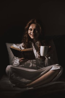 demonic smiling woman in nightgown holding bible and cup on bed clipart