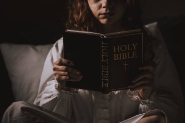 cropped view of creepy girl in nightgown reading bible on bed clipart