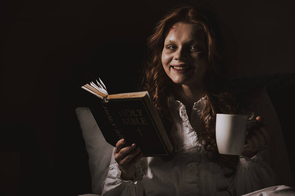 demonic smiling girl in nightgown holding bible and cup on bed