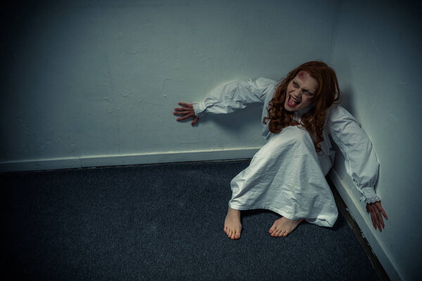 одержимая демоническая девушка в ночной рубашке кричит и сидит у стены
