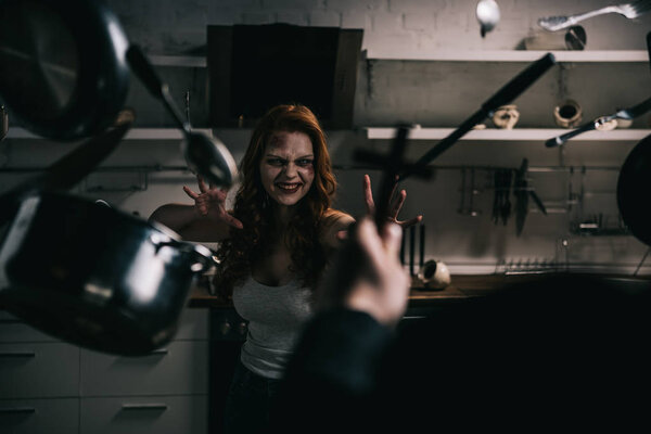 selective focus of demon with levitating cookware and exorcist with cross in kitchen