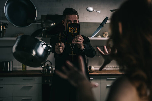 селективный фокус демона с левитационной посудой и экзорцист с крестом и библией на кухне
