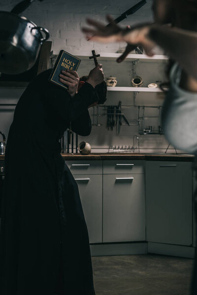 selective focus of evil demon with levitating cookware and exorcist with cross and bible in kitchen