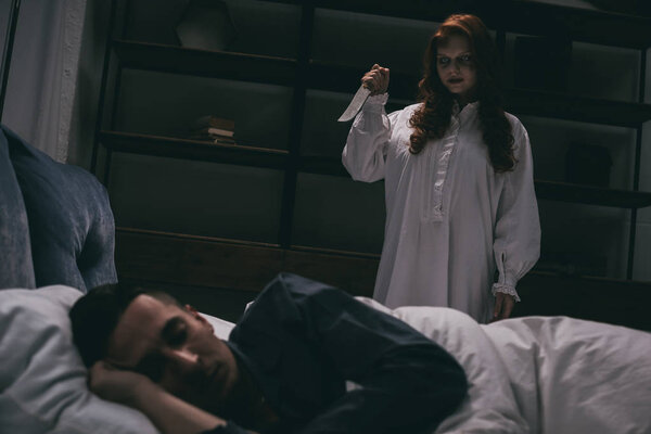 demoniacal creepy girl with knife standing over sleeping man in bedroom 