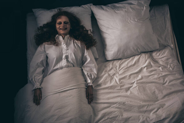 top view of creepy smiling demon in nightgown lying in bed