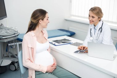 smiling doctor showing ultrasound scan to young pregnant woman in clinic clipart