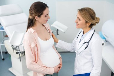 Genç gülümseyen hamile kadın klinikte jinekologla görüşüyor.