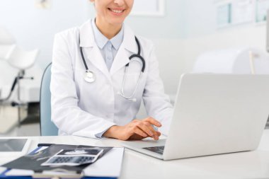 cropped view of positive doctor working on laptop in clinic with ultrasound scans on table clipart