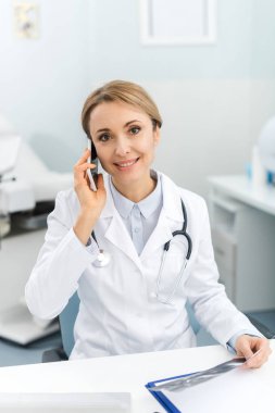 Gülümseyen doktor ultrason taraması yapıyor ve klinikte akıllı telefondan konuşuyor. 