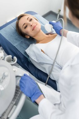 Genç doktor, klinikte ultrason taramasıyla gülümseyen hastanın tiroidini inceliyor.