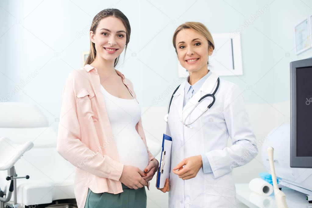 young pregnant woman standing with smiling gynecologist in clinic