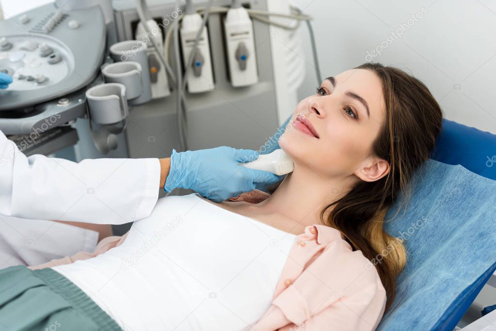 professional doctor examining thyroid of smiling patient with ultrasound scan in clinic