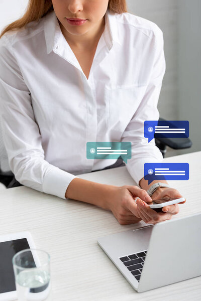 Cropped view of businesswoman holding smartphone near gadgets and glass of water on table, speech bubbles illustration