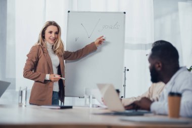 selective focus of smiling businesswoman pointing with finger at goals lettering and talking with african american colleague clipart