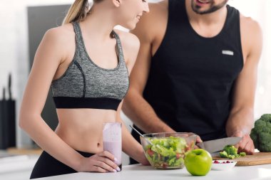 Mutfakta yemek pişiren adamın yanında elinde bir bardak smoothie tutan neşeli kadın manzarası. 