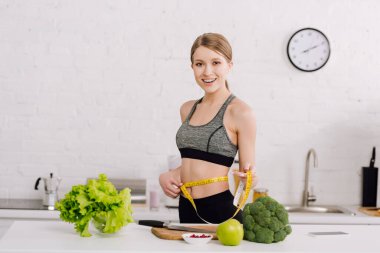 sportive girl measuring waist near fresh food in kitchen  clipart