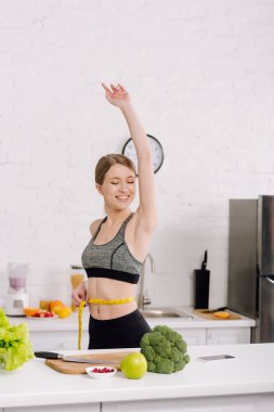 cheerful girl measuring waist near fresh food in kitchen  clipart