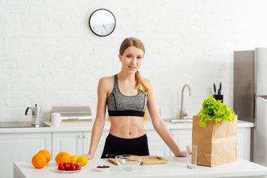 Spor kıyafetleri içinde mutlu bir kadın kese kağıdının yanında ve mutfakta meyveler 