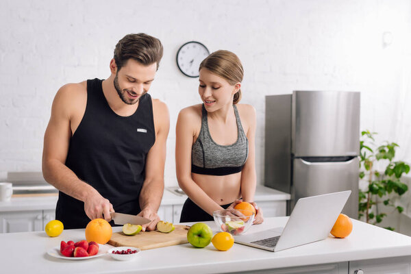 happy man cutting apple near girl in sportswear and laptop 