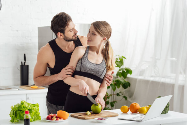 happy girl cutting apple near sportive man, laptop and fruits in kitchen 