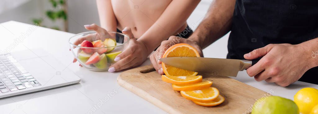 panoramic shot of man cutting orange near woman, salad and laptop 