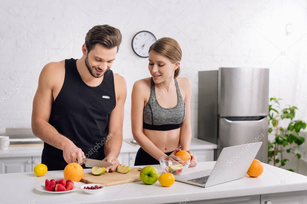 happy man cutting apple near girl in sportswear and laptop 