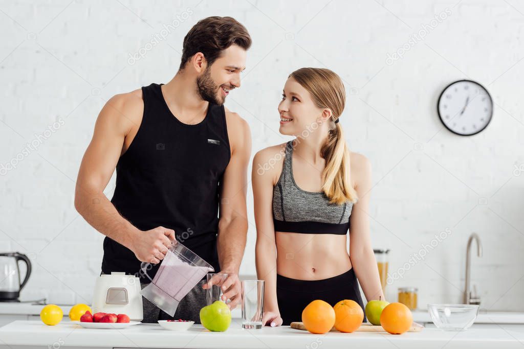 sportive man pouring smoothie in glass near happy woman and fruits 