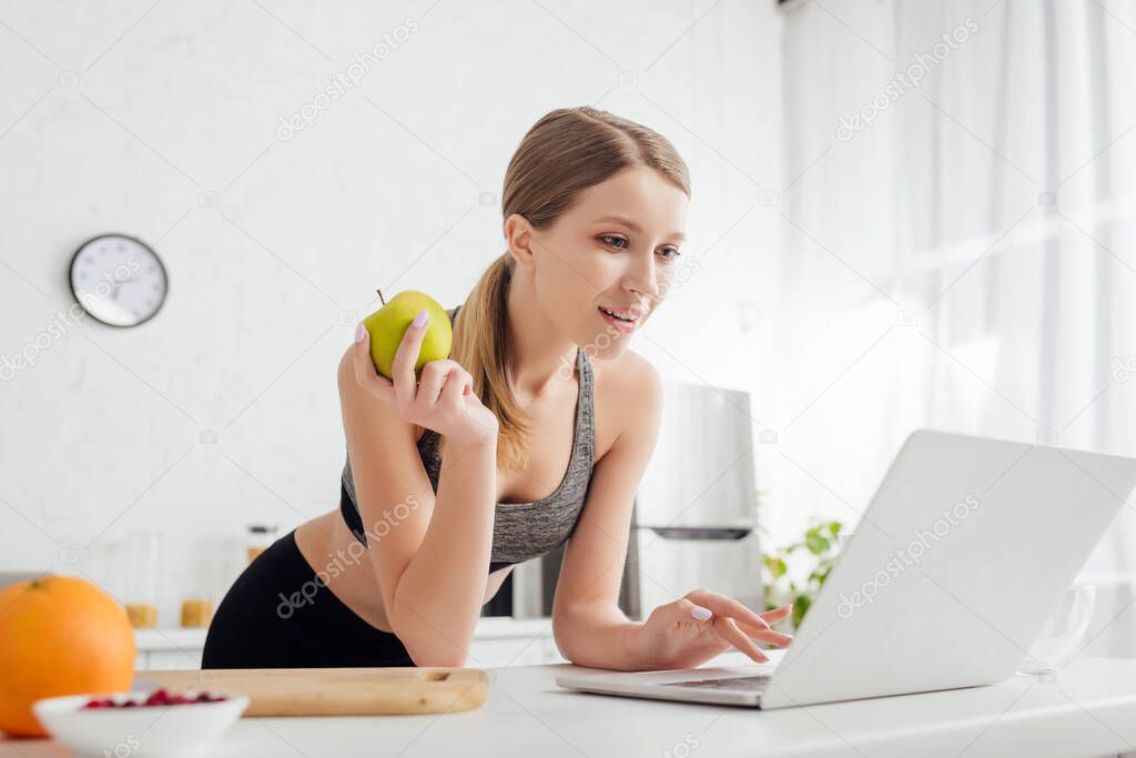selective focus of sportive woman holding tasty apple and using laptop 