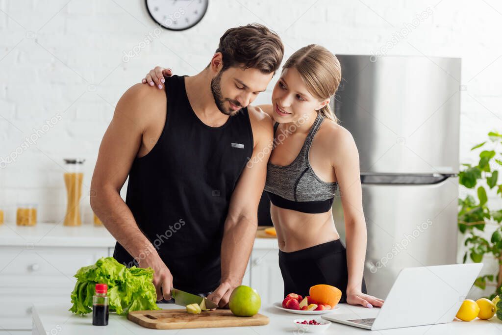 attractive girl looking at man cutting apple near laptop and fruits in kitchen 