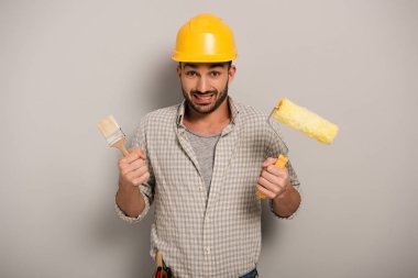 confused manual worker in hardhat holding paint roller and brush on grey  clipart