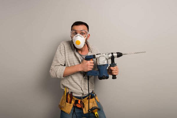 scared manual worker in safety mask holding electric drill on grey