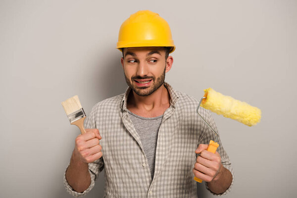thoughtful manual worker in hardhat looking at paint roller and brush on grey 