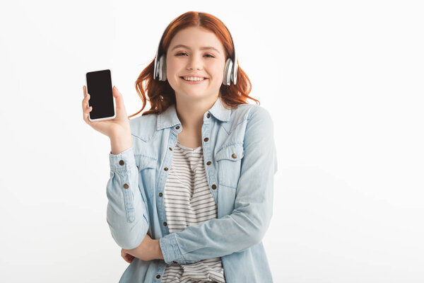 cheerful teen girl listening music with headphones and showing smartphone with blank screen, isolated on white