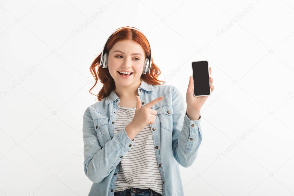 excited teenager listening music with headphones and pointing at smartphone with blank screen, isolated on white 