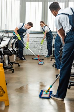 multicultural team of young cleaners washing floor with mops in office clipart