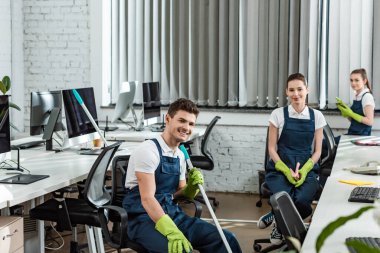Gülümseyen temizlik ekibi modern ofiste oturuyor ve kameraya bakıyor.