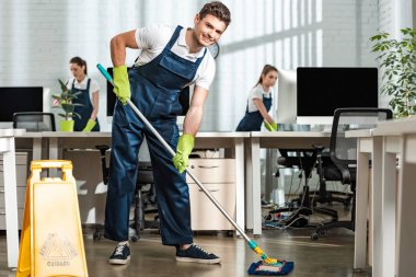 young cleaner looking at camera while washing floor with mop in modern office clipart