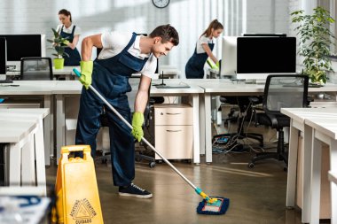 young, smiling cleaner washing floor with mop in modern office clipart